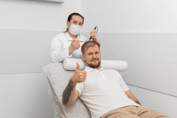 Pacjent w Turkus Hair Transplant Clinic podczas zabiegu na skórze głowy: specjalistka w masce i rękawiczkach wykonuje iniekcję, pacjent gestem kciuka w górę sygnalizuje komfort.